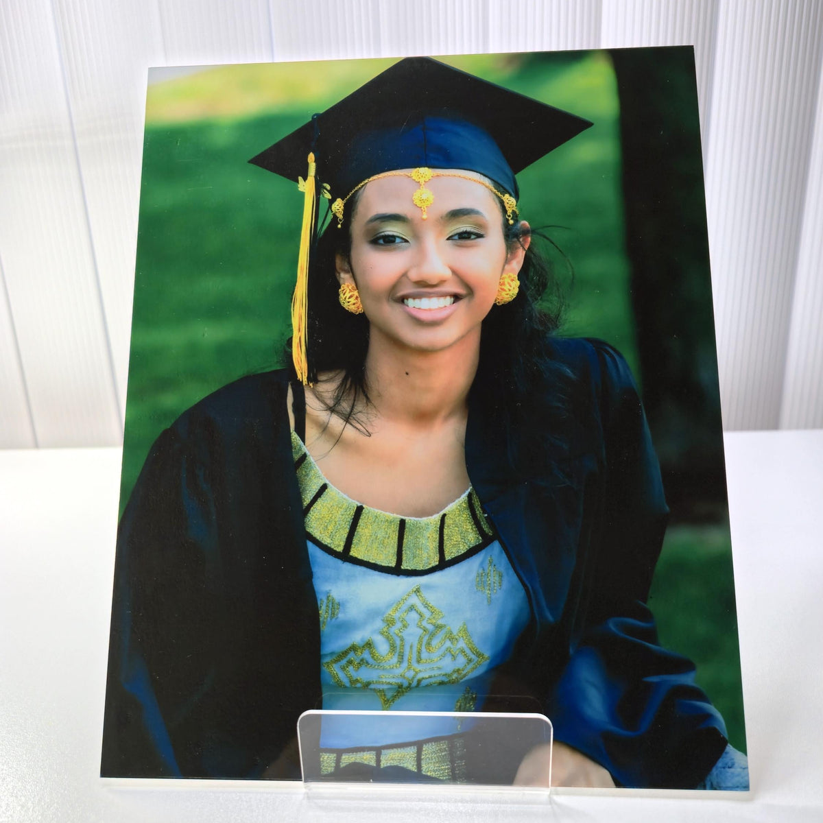 A printed portrait photograph of a smiling woman in a graduation cap and gown, displayed upright on a clear acrylic stand against a white paneled background.