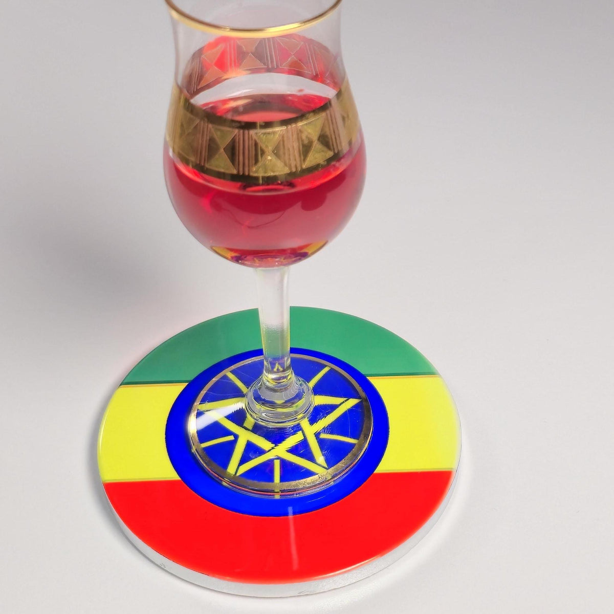 A clear stemmed glass filled with red liquid sits on a round coaster printed with the Ethiopian flag colors and a yellow star emblem in the center, photographed on a light gray background.