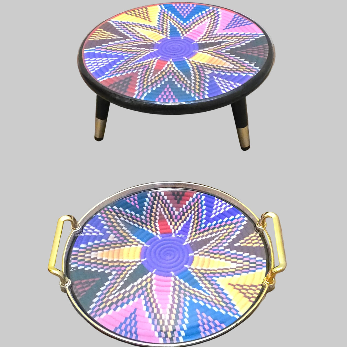 Two decorative round trays with colorful geometric starburst patterns, one with black legs and gold trim, and one with gold handles and a gold rim, displayed on a wooden floor against a white wall.