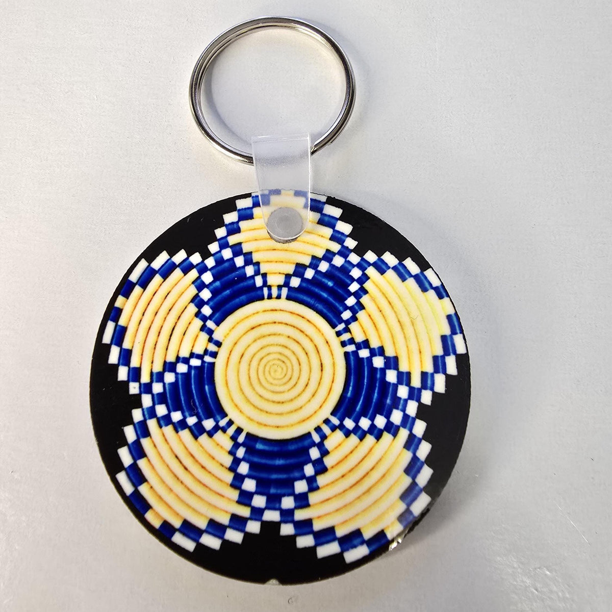A round keychain featuring a colorful geometric pattern with concentric circles in yellow, blue, and white, set against a black background. The keychain has a silver metal split ring and a clear plastic connector.