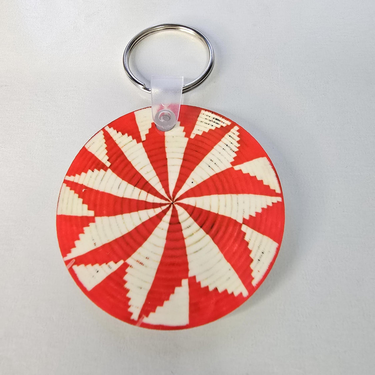 A circular woven keychain with a red and off-white geometric spiral pattern, attached to a silver metal split ring.