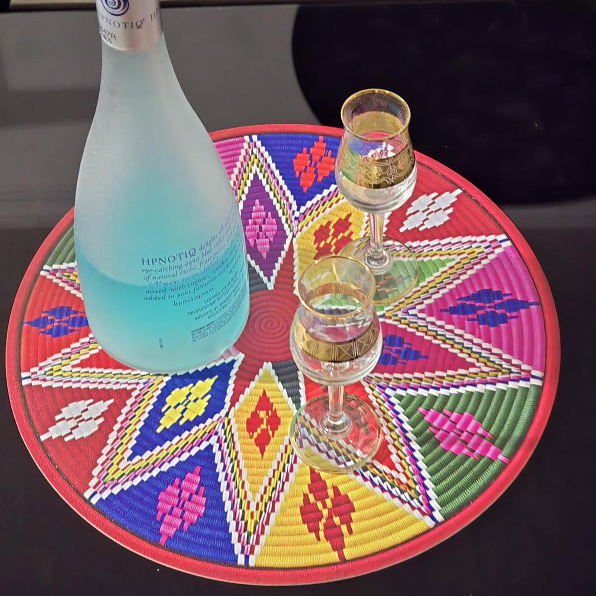 A bottle of Hipólito Rosado sparkling wine with two small glasses, set on a colorful circular woven placemat on a dark reflective table.