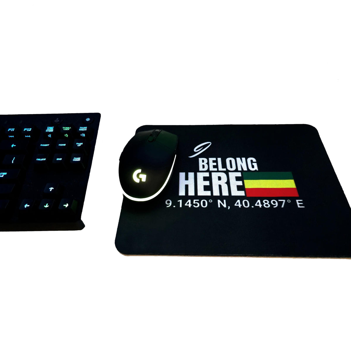 A black computer mouse pad with a white 'BELONG HERE' graphic, coordinates, and a small green-yellow-red flag, shown alongside a black gaming mouse and part of a black keyboard on a white background.