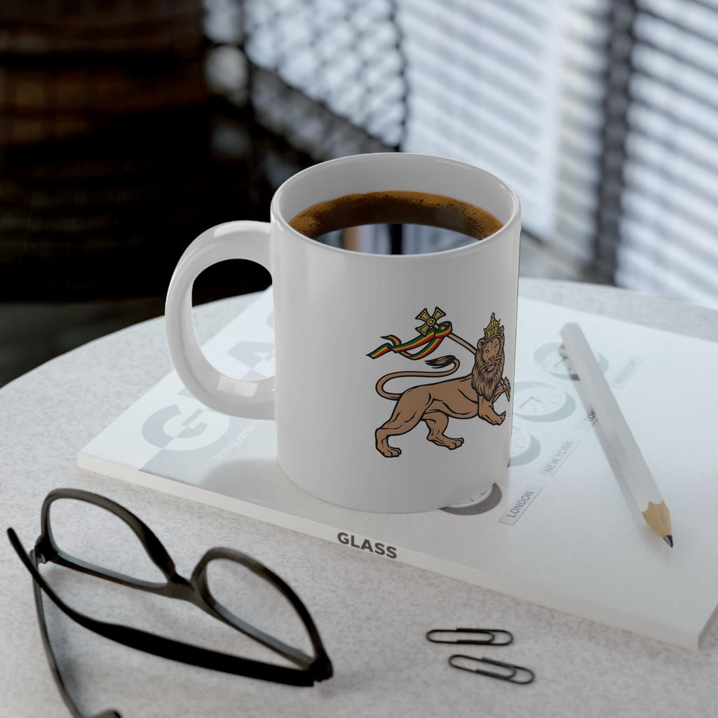 A white ceramic coffee mug with a printed lion graphic sits on a tabletop with a cup of coffee inside. The mug rests on a book or folder, with a pencil, eyeglasses, and paper clips nearby in a real indoor setting with window blinds in the background.