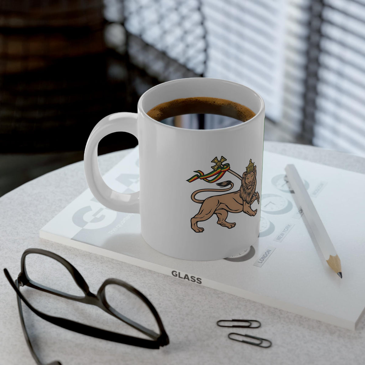 A white ceramic coffee mug with a printed lion graphic sits on a tabletop with a cup of coffee inside. The mug rests on a book or folder, with a pencil, eyeglasses, and paper clips nearby in a real indoor setting with window blinds in the background.