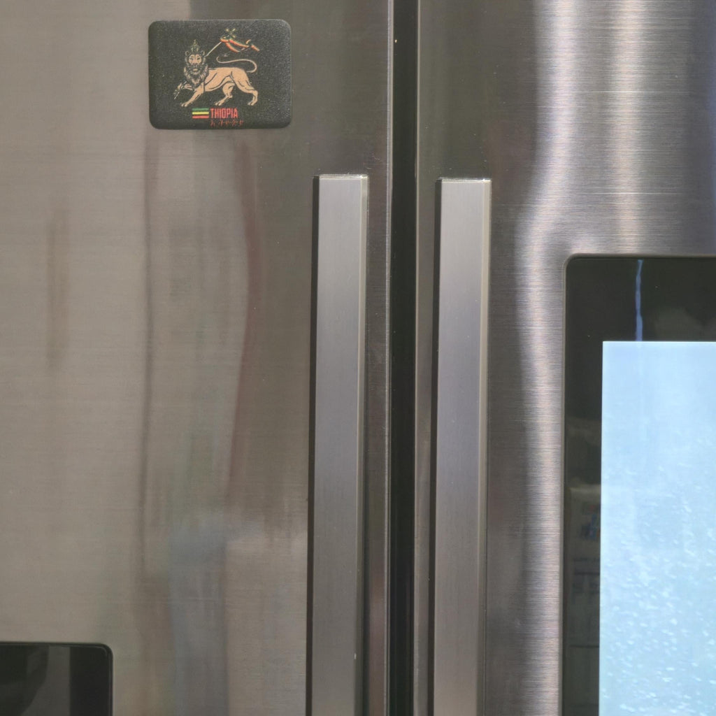 Close-up of a stainless steel refrigerator door with two vertical handles, a small black square logo sticker near the top left, and part of a blue-lit control panel visible on the right.