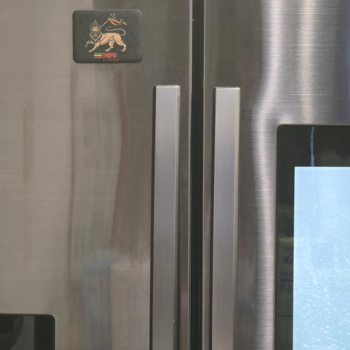 Close-up of a stainless steel refrigerator door with two vertical handles, a small black square logo sticker near the top left, and part of a blue-lit control panel visible on the right.
