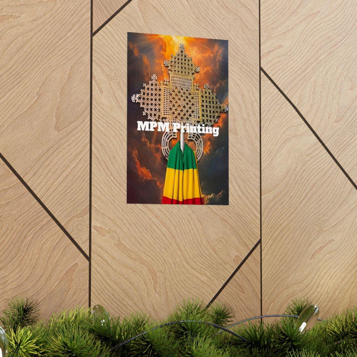 A vertical art print featuring a white geometric cross on a fiery orange and yellow background, with a green, yellow, and red striped flag at the bottom and the text 'MPM Printing' across the center. The print is mounted on a light wood paneled wall above a green pine garland.