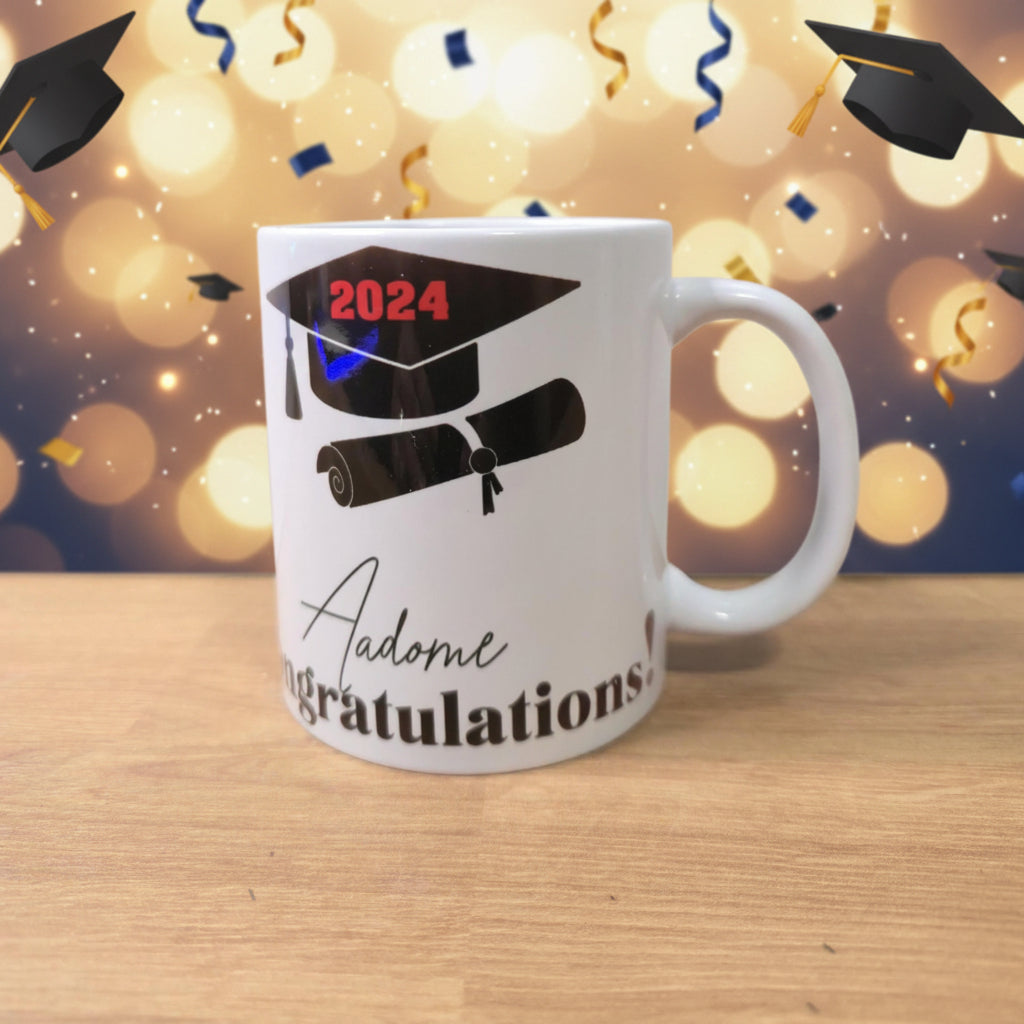 A white ceramic graduation mug sitting on a light wooden table against a plain light gray wall. The mug features a black graphic of a graduation cap and diploma scroll with '2024' printed in red on the cap, and the words 'Adome Congratulations' in black script and serif fonts below.