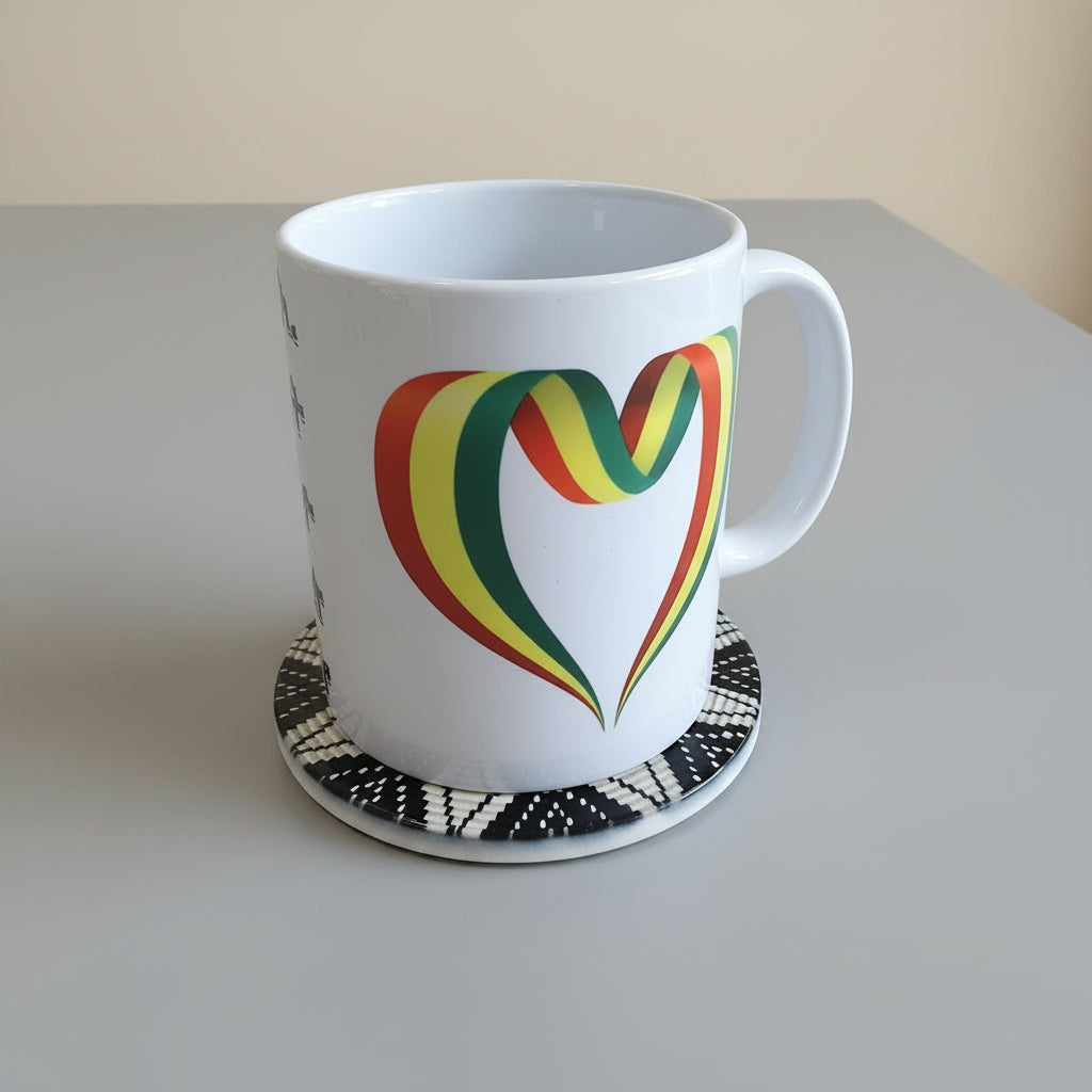 A white ceramic mug with a colorful heart-shaped graphic in red, yellow, green, and black, sitting on a black and white patterned coaster against a neutral beige background.