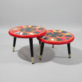 Two round nesting side tables with red rims and colorful geometric woven tops, shown on a black marble surface against a gray wall. White arrows and text indicate table heights of 10 inches and 6 inches.