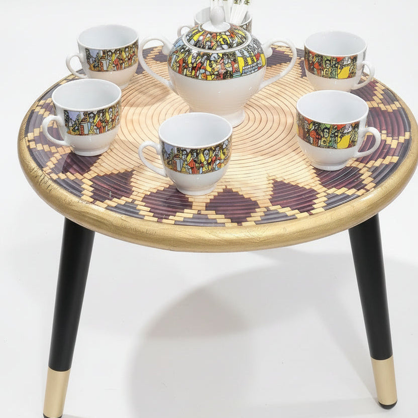 A ceramic tea or coffee set arranged on a round woven tray, including a lidded teapot, sugar bowl, and four matching cups, with white spoons in the teapot. The cups and teapot have a colorful illustrated band around the middle, while the rest of the set is white.