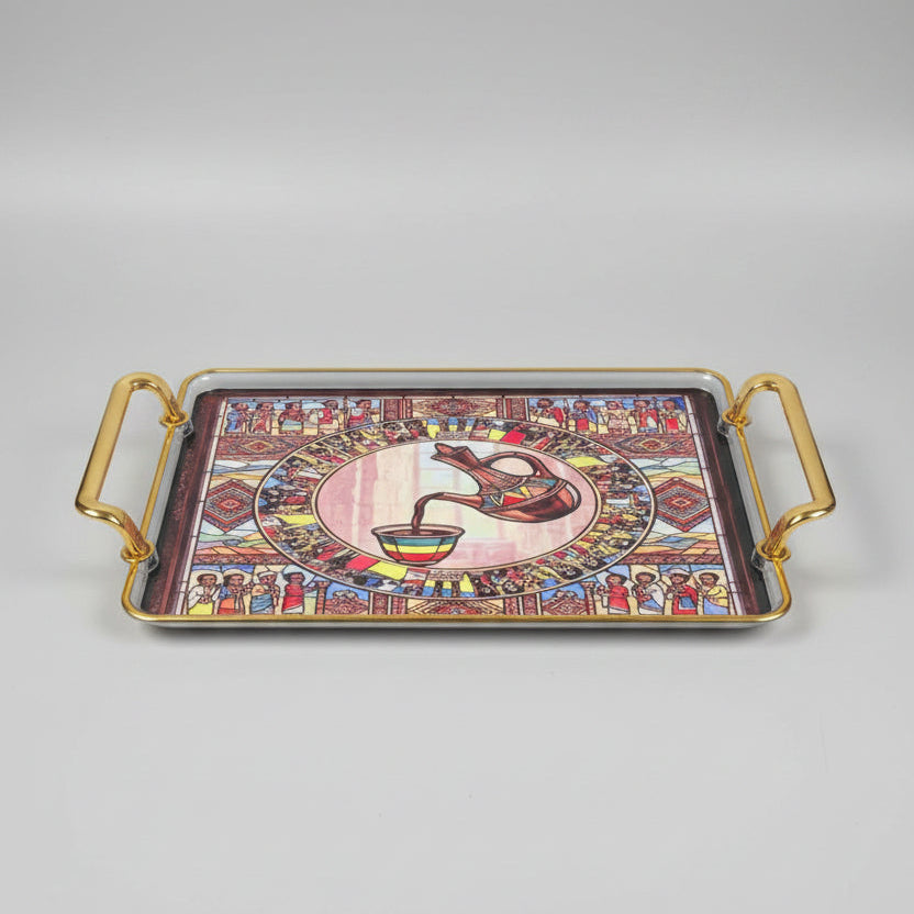 A rectangular decorative tray with gold-toned handles and a colorful printed design featuring a central coffee pot pouring into a cup, surrounded by illustrated figures and geometric patterns, placed on a black marble surface.