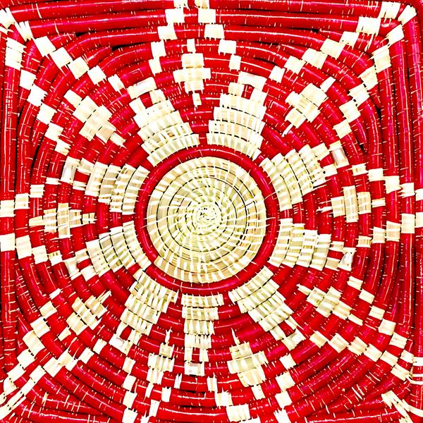 A top-down view of a hand-woven basket or tray with a geometric radial pattern in red and natural beige fibers.