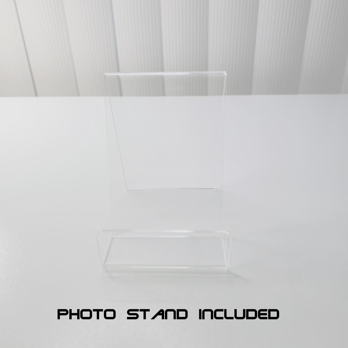 A clear acrylic photo stand with a rectangular base and a smaller upright rectangular piece, shown on a white surface against a white ribbed background. Black text at the bottom reads 'PHOTO STAND INCLUDED'.