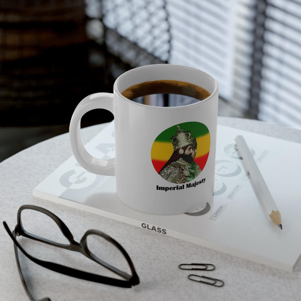 A white ceramic coffee mug filled with black coffee sits on a light gray tabletop, styled with eyeglasses, a pencil, paper clips, and a book. The mug features a circular graphic of a bearded man in military armor with the text 'Imperial Majesty' and the colors green, yellow, and red.